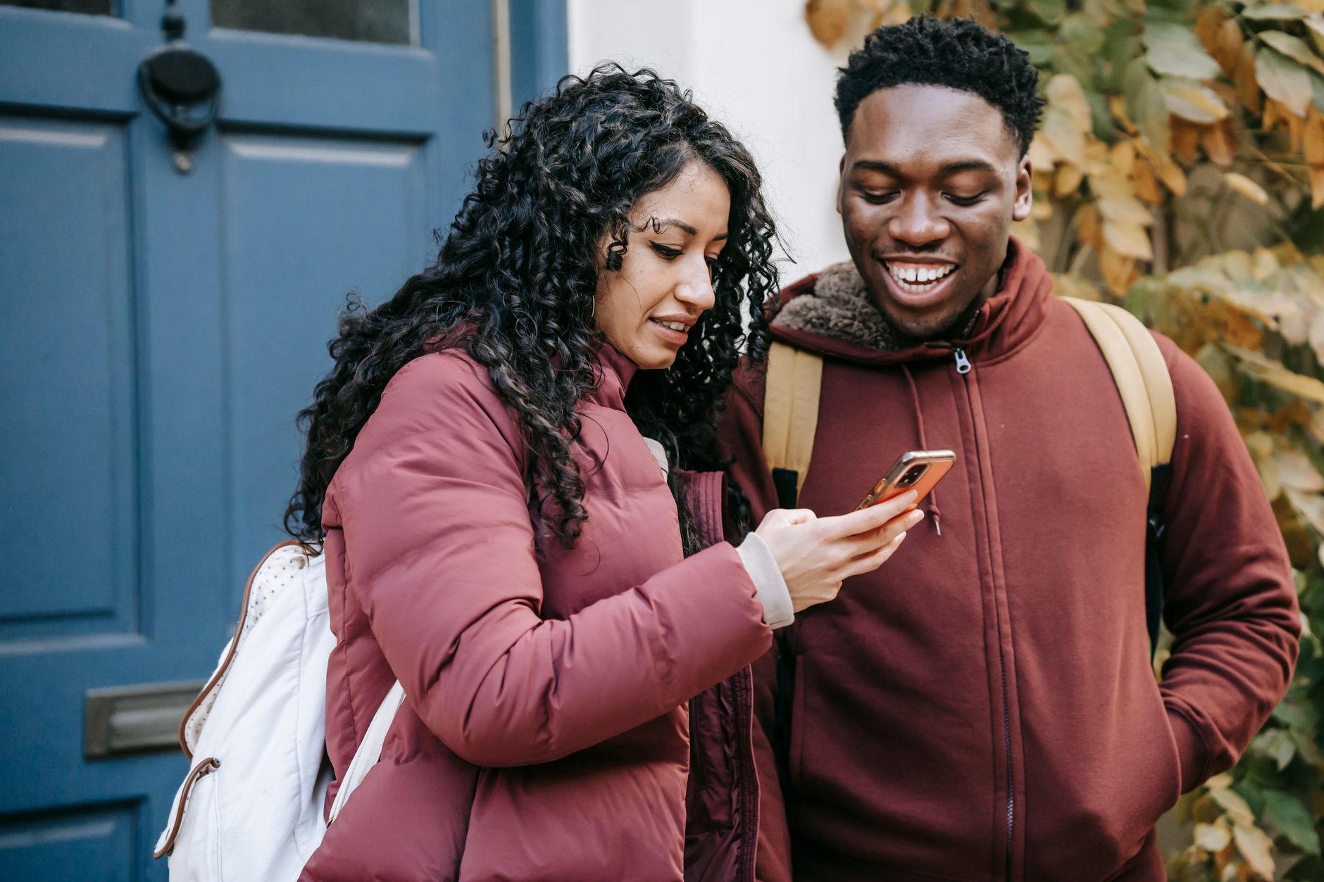 Couple diversifié et joyeux en vêtements chauds utilisant un téléphone portable près de la porte d'un bâtiment dans la rue.