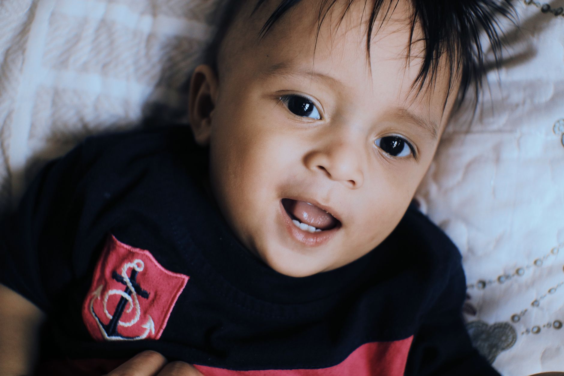 Jeune enfant allongé sur un tissu clair, bouche entrouverte, vêtu d’un haut bleu marine avec un écusson rouge à motif d’ancre.