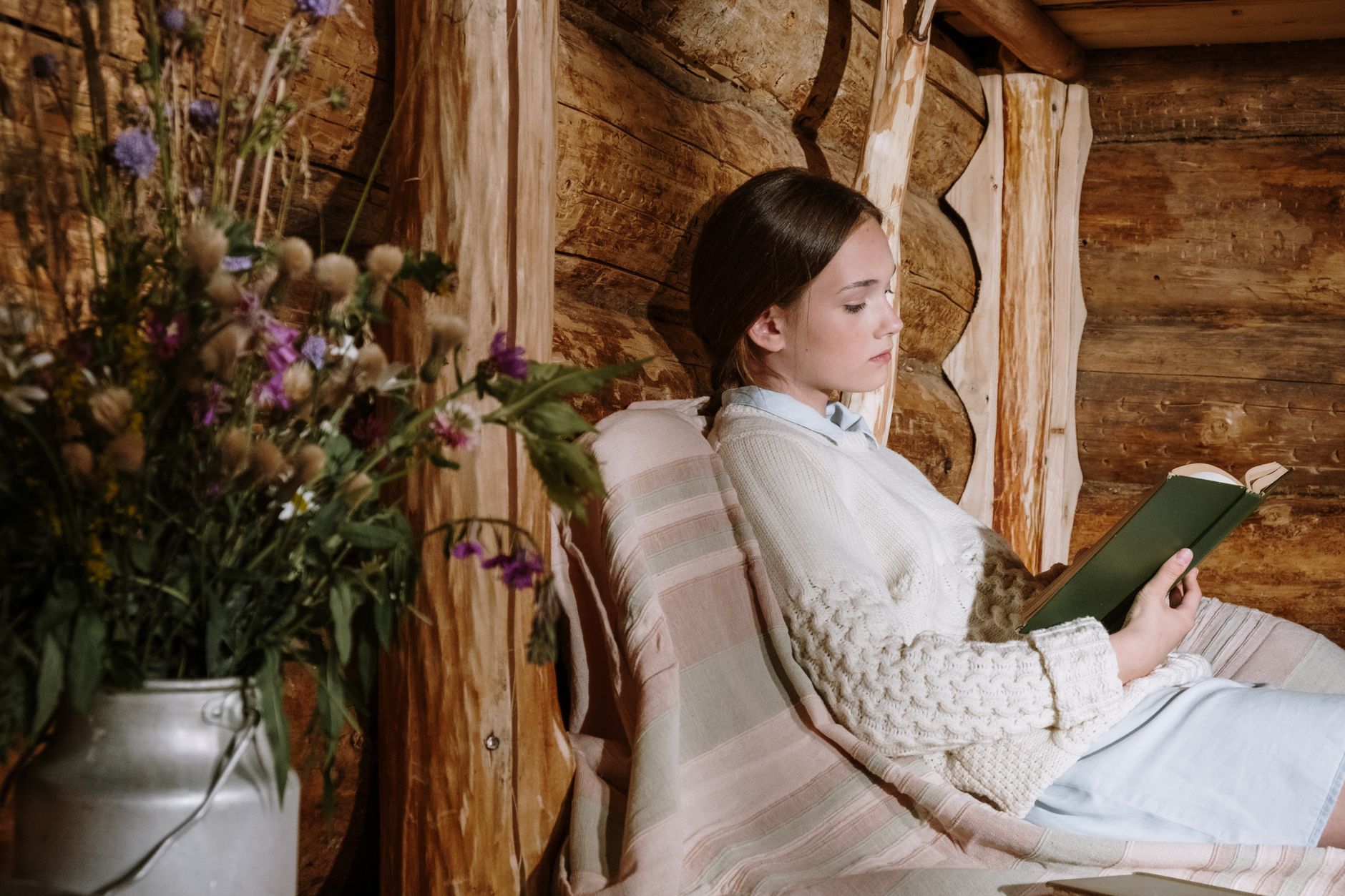 Jeune femme lisant un livre, adossée à une couverture rayée dans une pièce en rondins, avec un bouquet de fleurs sauvages au premier plan.