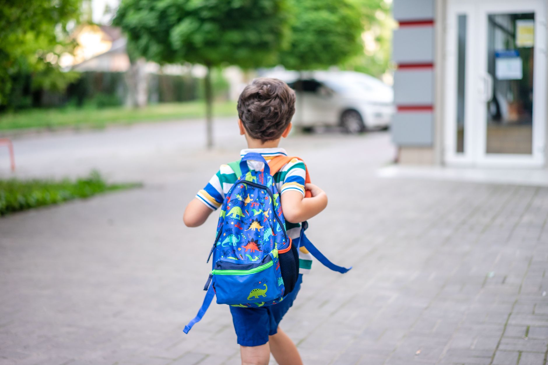 Petit garçon vu de dos marchant sur un trottoir pavé avec un sac à dos bleu à motifs de dinosaures, près d’un bâtiment à porte vitrée.
