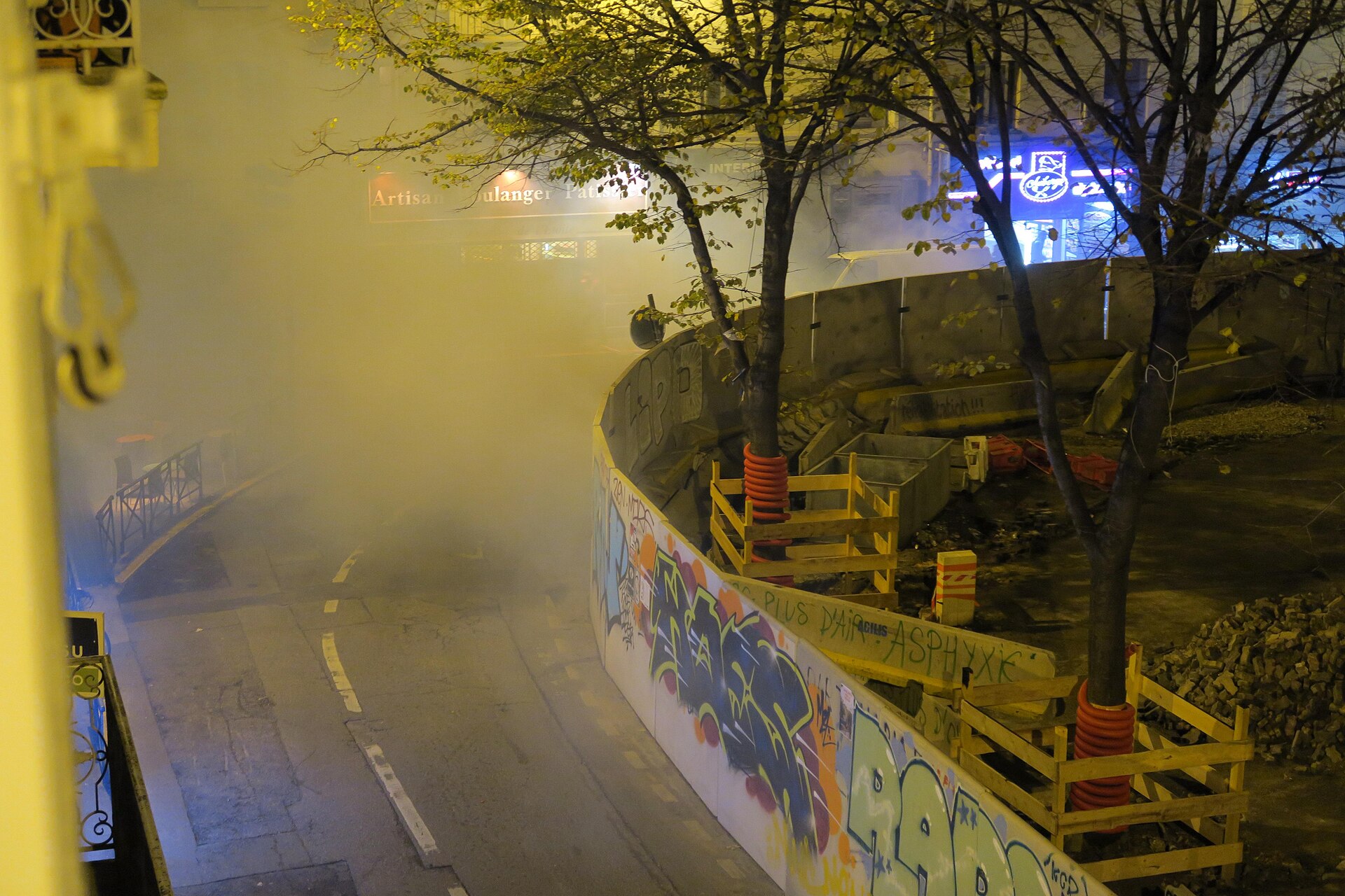 Rue de nuit envahie par un nuage de gaz lacrymogène, le long d'une palissade de chantier couverte de graffitis