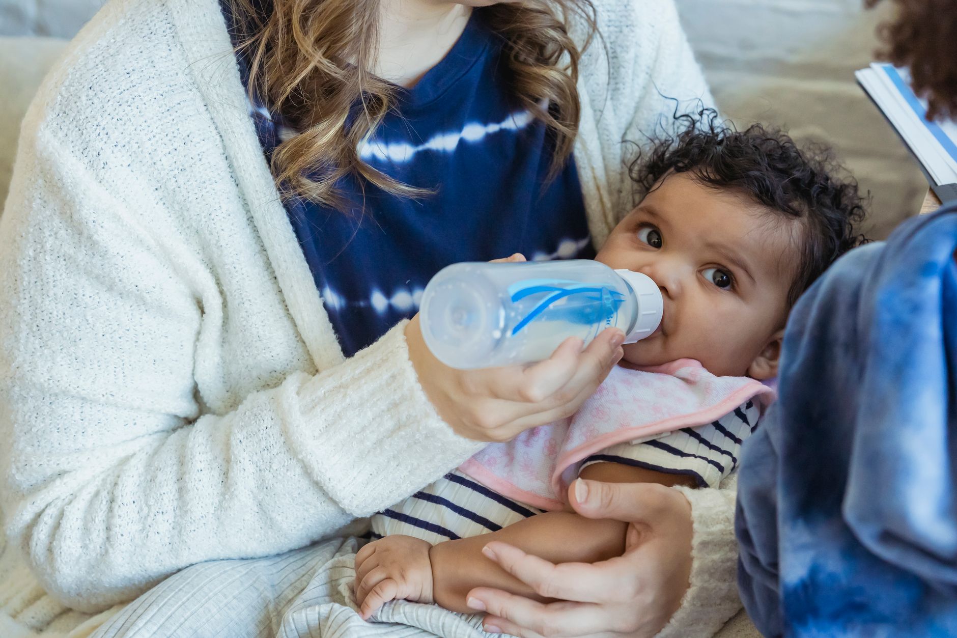 Un adulte tient un bébé dans ses bras et lui donne un biberon transparent, assis sur un canapé clair dans un intérieur.