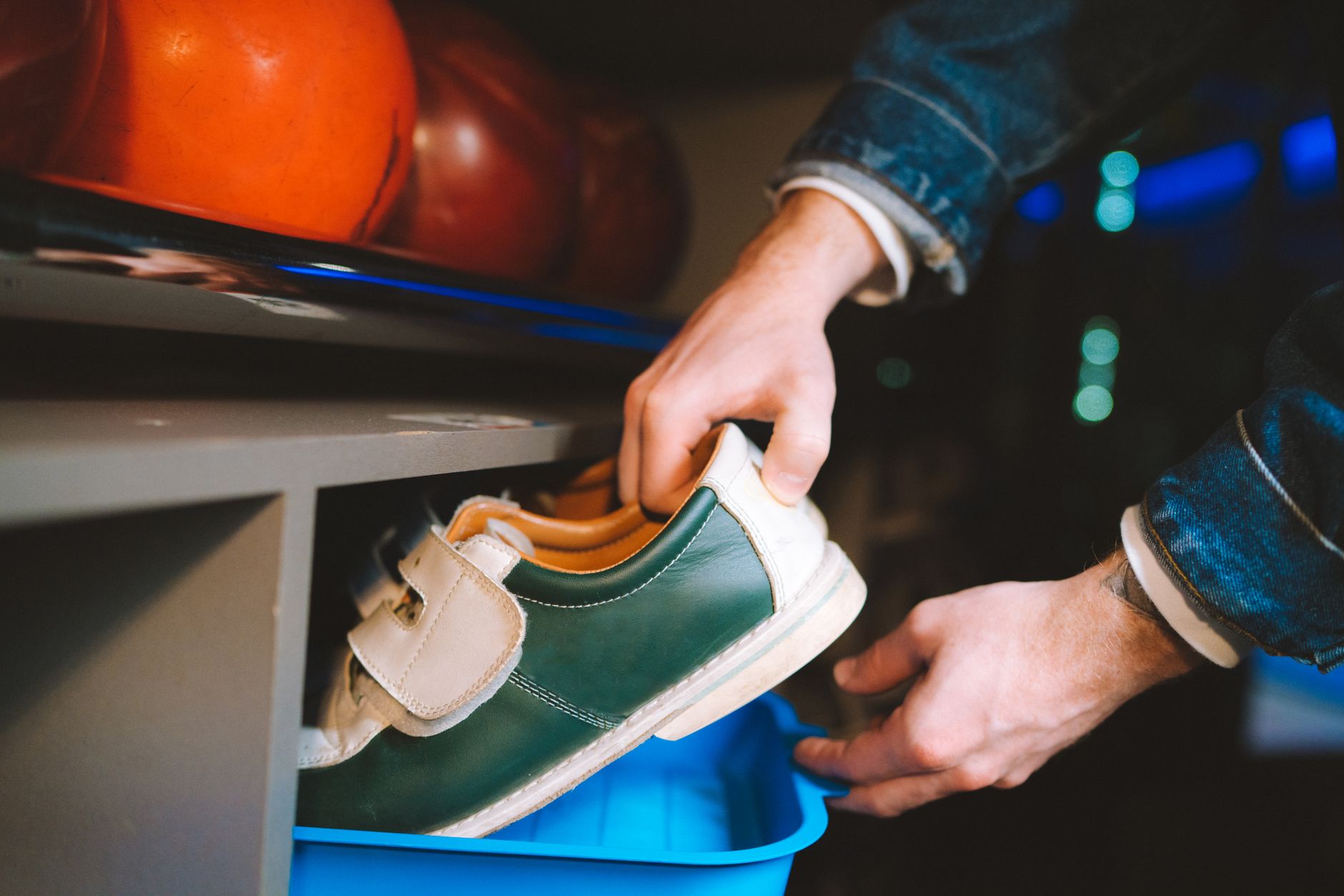 Une personne prend une chaussure de bowling verte et blanche à scratch dans un casier, sous des boules de bowling orange et rouges.